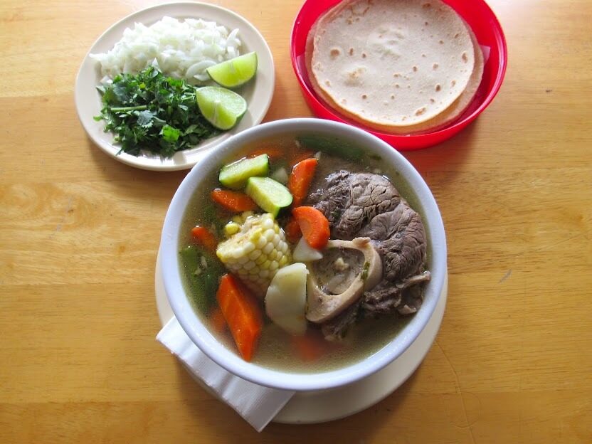A bowl of caldo de res — beef, corn, carrot and zucchini in the broth — with onion, cilantro and warm tortillas alongside